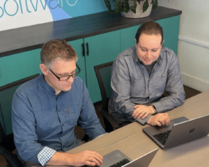 Two professionals working on laptops at a desk with the '303 Software' logo in the background, indicating a collaborative environment at a Colorado software and app development company.